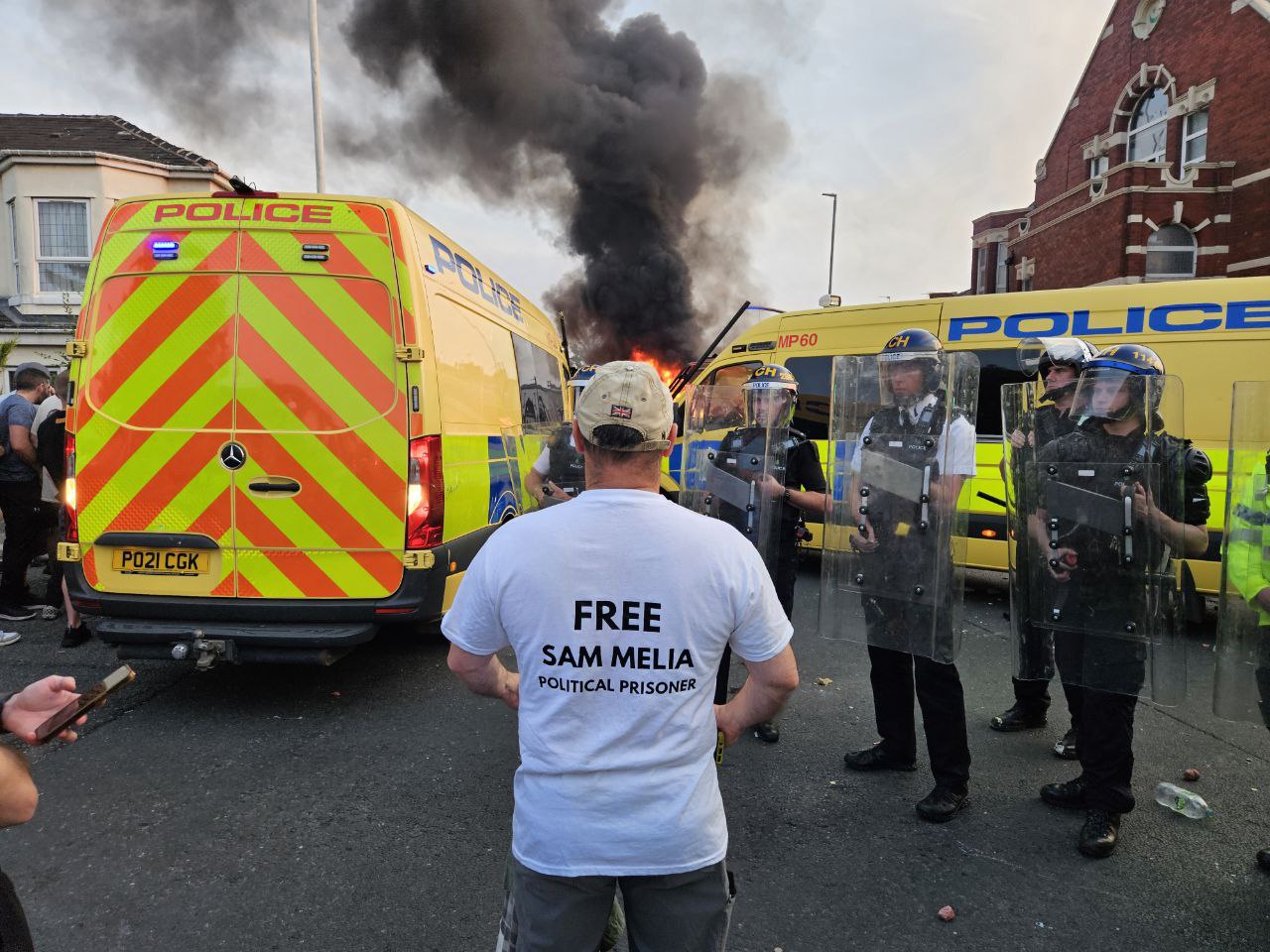 Miles facing down riot police, wearing a t-shirt featuring the slogan 'Free Sam Melia'